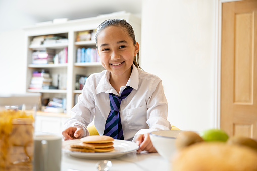 Waist-up view of multiracial 11 year old girl in uniform sitting at dining table with plate of pancakes and smiling at camera.