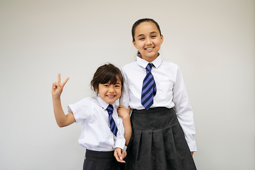 Front view of 6 and 11 year old girls standing side by side, grinning at camera, and gesturing peace sign against white background.