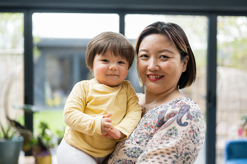 Close view of multiracial woman in early 40s holding toddler son inside family home, both in casual clothing, and smiling at camera.