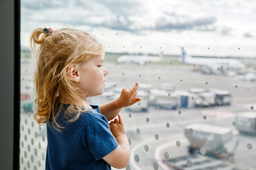 Cute little toddler girl at the airport, traveling. Happy healthy child waiting near window and watching airplanes. Family going on summer vacations by plane