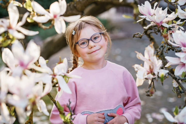 花のモクレンの木の下に眼鏡をかけたかわいい春のファッション幼稚園の女の子。小さな幸せな子供と春。 - sunlight flower magnolia flower head ストックフォトと画像