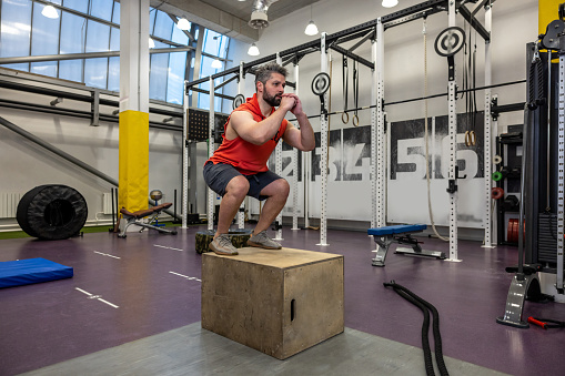 Caucasian bearded man having workout, jumping up on wooden box in gym gym.