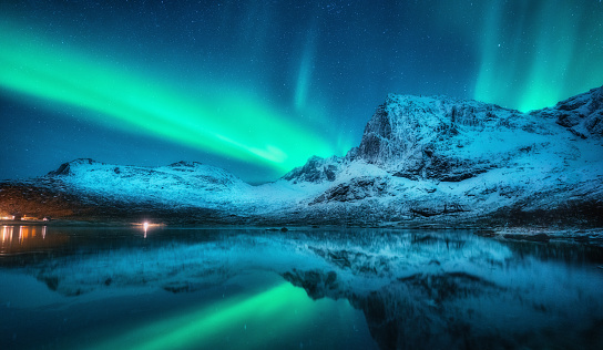 Northern lights over the snowy mountains, sea, reflection in water at winter night in Lofoten, Norway. Aurora borealis and snow covered rocks. Scenery with polar lights, sky with stars and fjord