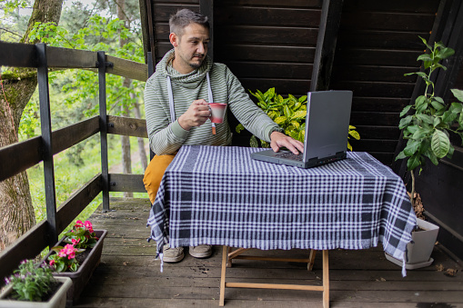Young businessman is working on his laptop on the balcony of his country house. He is drinking his first morning coffee.
Rural scene with modern technology.