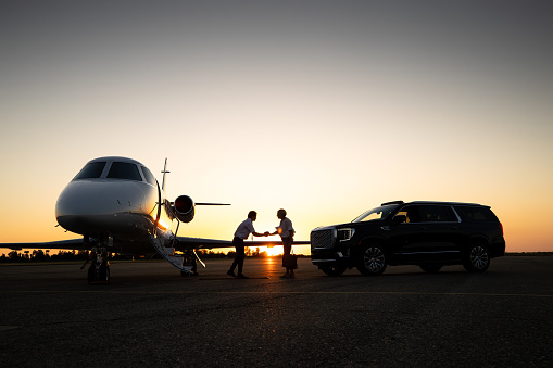 A young Asian pilot shakes hands with a mature White woman. A private jet and a luxury SUV parked next to them.