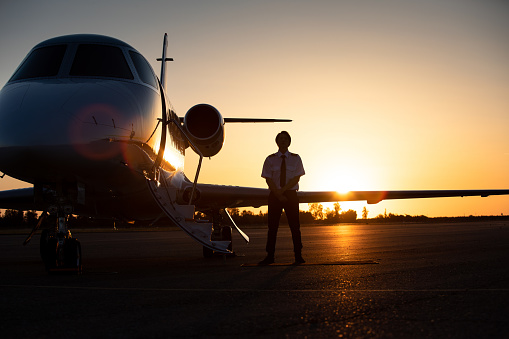 A young Asian pilot standing next to a private jet at dusk.