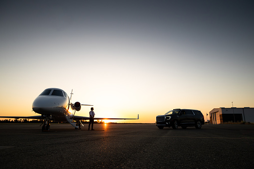 A young Asian pilot stands amongst a private jet and a black luxury SUV at sunset.