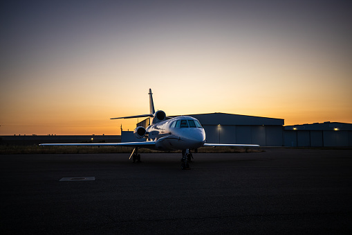 A private plane sits parked on a runway with an airplane hangar in the background.