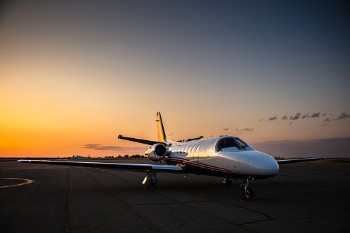 A private plane sits parked on a runway with the sun setting in the background.