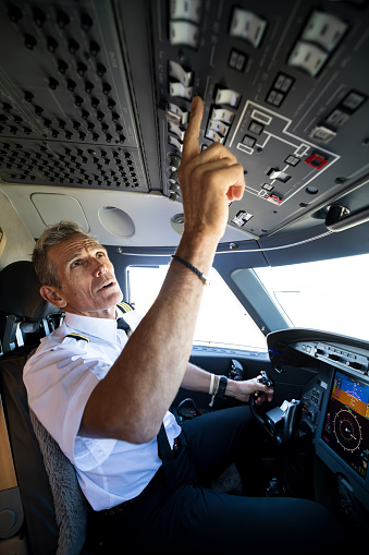A mature White pilot pressing buttons inside the cockpit of a private jet.