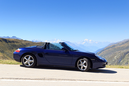 Realp, Switzerland - August 13, 2022: Blue roadster Porsche Boxster 986 and panorama with mountains Dom and Weisshorn at Furka Pass road. The car is a mid-engine sports car manufactured by Porsche.