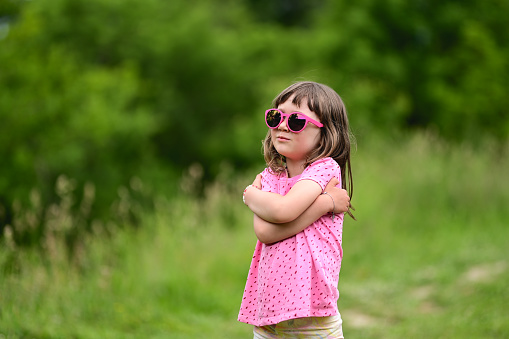 serious preschool girl in a pink sunglasses standing in a green meadow.