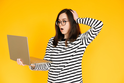Picture of a cute impressionable lady in a striped T-shirt with glasses, reading the news or writing on a modern laptop device, hand head isolated yellow background color.