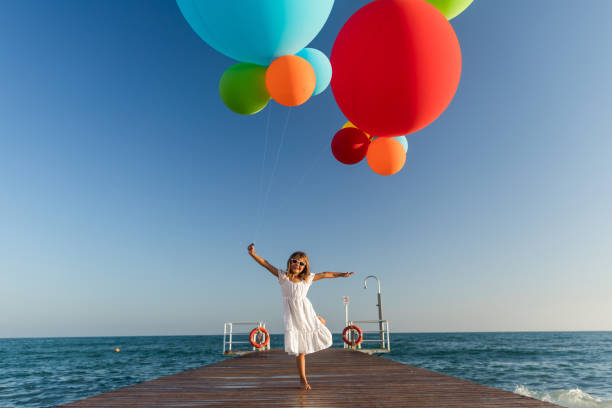 kleines mädchen im weißen kleid läuft auf hölzernem kai mit farbigen luftballons auf das meer zu - balloon child people color image stock-fotos und bilder