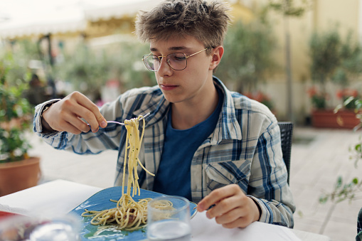 Teenage boy eating spaghetti with mussels. Sidewalk restaurant.\nCanon R5