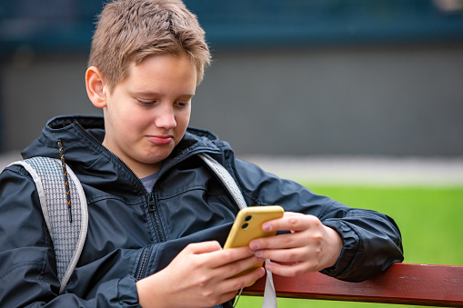 A young smiling teenage school boy sitting  outdoors on the bench looking on his mobile phone and watching funny video online at school break. Boy with dark blond hair and blue eyes wearing black jacket and sitting outdoor in the park in front of a modern building holding his yellow mobile phone.  School boy with backpack.
Background is modern building out of focus.