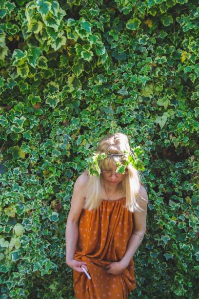 A blond woman with ivy leaves in her hair and an orange dress is standing in front of an ivy wall