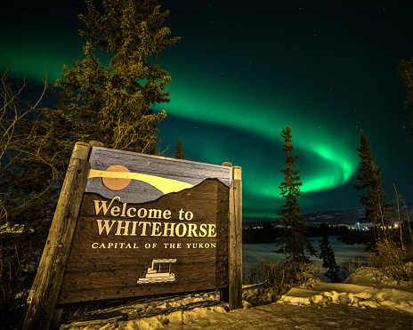 Welcome to Whitehorse sign in Yukon Territory with bright green aurora borealis northern lights wilderness city of arctic area.