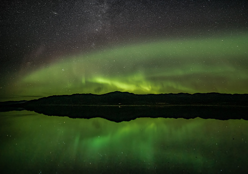 Ghostly northern lights scene in Yukon, Canada during fall with spectacular stars and green aurora borealis band across the northern landscape. Incredible reflection of green bands in the water below.