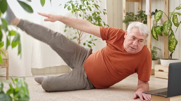 Happy mature senior man doing exercises of gymnastics at home online with laptop. Healthy lifestyle, fitness, recreation, well being. Elderly male exercising training, stretching. Old man working out.