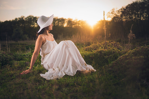 Young woman posing sitting in forest at sunset. Back view in white dress