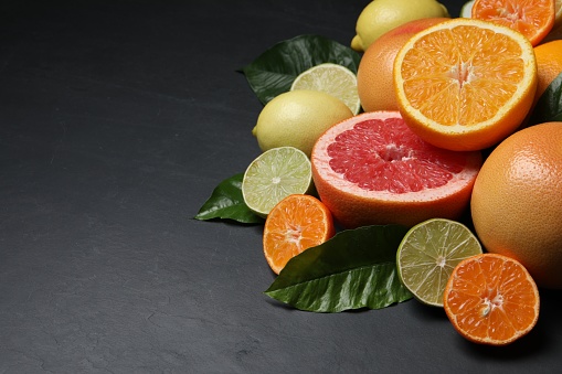 Different ripe citrus fruits with green leaves on black table. Space for text