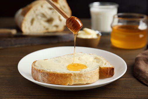 Pouring honey from dipper onto sandwich with butter on wooden table, closeup