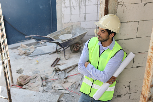 Caucasian technician civil engineer or specialist inspector standing and crossed arms while holding blueprint and looking away after project complete at Industrial building site. Construction concept