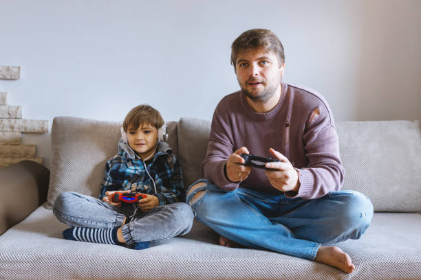 istock Father And Son Sitting On Sofa In Lounge Playing Video Game 1495237614