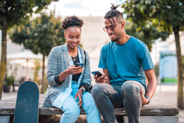 istock Cute Young Black Couple Sitting On A Bech Outdoors, Using Their Smart Phones 1495238084