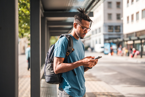 Young Black male using a smart phone, trading cryptocurrency and surfing the net. He is outdoors, commuting in the city on a sunny summer day.