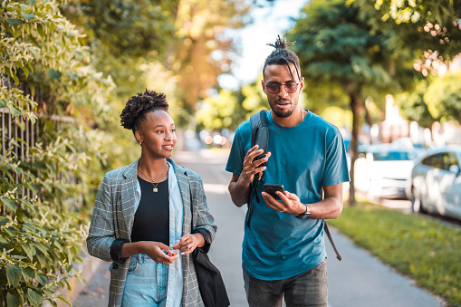 Beautiful young African American couple walking in the city, scrolling on their smart phones and hanging out. They are wearing casual clothing.