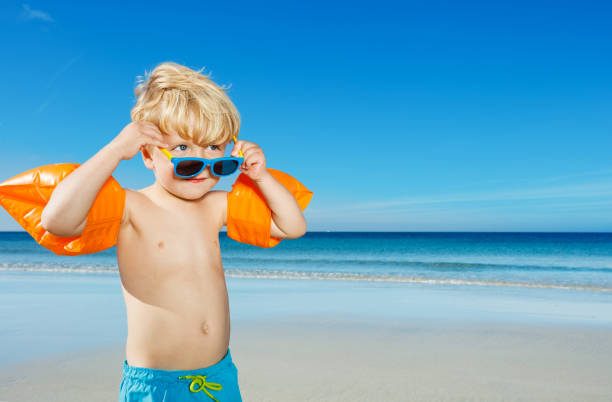istock Boy in inflatable shoulder straps holding sunglasses on beach 1495246550