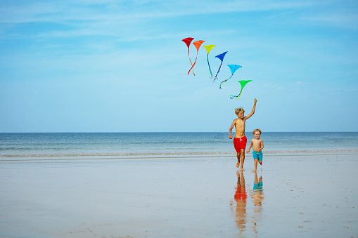 Two children, boys run holding many colorful kite at the beach smiling over the sky during vacation