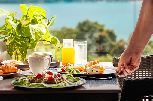 Waiter serving table with drinks and food in restaurant in luxury resort with sea view. Closeup of food stylish. Restaurant serving. Healthy breakfast in hotel. Summer vacation.