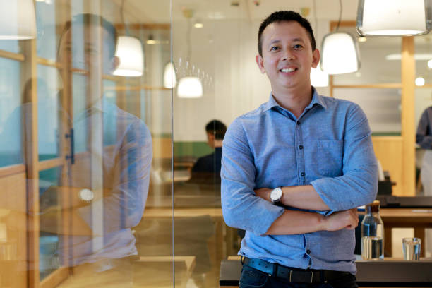 A portrait captures a young, confident Asian man standing with his arms folded and a bright smile on his face. He leans against his office desk, radiating positivity.
