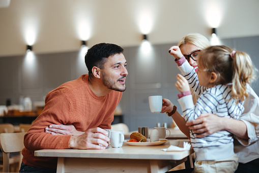 Handsome smiling man is leaning over the table while his daughter is playfully showing him something. Unrecognizable woman is holding her.