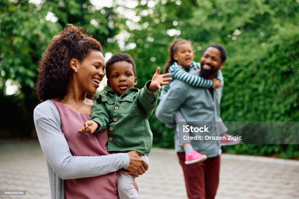 Let's go over there mommy! Happy African American woman holding her small son while walking outdoors. Father and daughter are in the background. Family Stock Photo