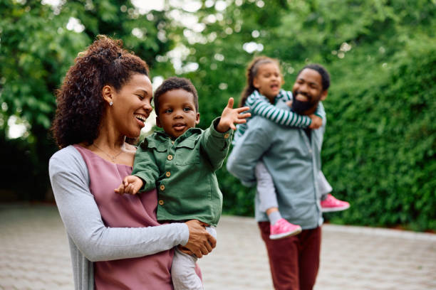 Let's go over there mommy! Happy African American woman holding her small son while walking outdoors. Father and daughter are in the background. african american stock pictures, royalty-free photos & images