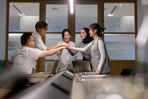 A multiethnic and diverse group of Asian colleagues join hands together in unity in a meeting room. They form a creative team of casual business coworkers, engaged in a project meeting at a modern office. Their collaboration embodies the spirit of startup teamwork.