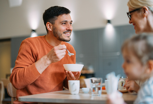 Close up shot of a cheerful handsome man sitting at the table and having breakfast with his unrecognizable wife and daughter. He is holding a bowl and a spoon while looking away.