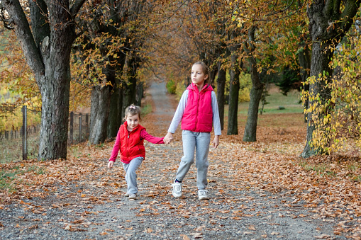 Full length of two happy girls holding hands and walking in autumn park