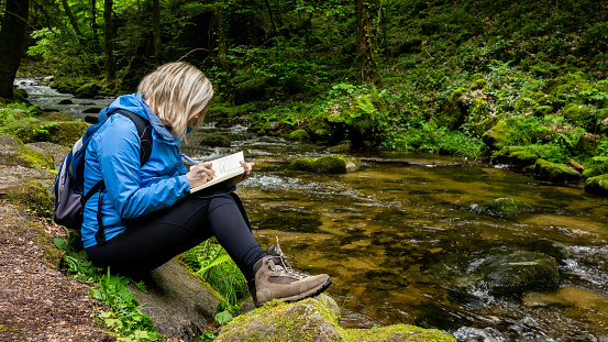 Young blond woman in a blue parka writing in her diary by a fresh forest creek in mountains. Travel alone. Black Forest.