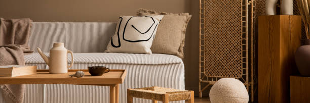 istock Warm and cozy living room interior with beige modular sofa, patterned pillow, brown plaid, wooden coffee table, books, stool, pitcher and personal accessories. Home decor. Template. 1495317917