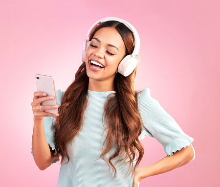 Phone, music headphones and woman singing in studio isolated on a pink background. Cellphone, radio singer and happy female with mobile streaming, laughing and listening to audio, sound or podcast.