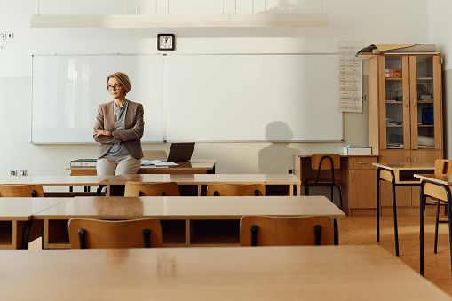 An elderly woman teacher writes with chalk on a blackboard.