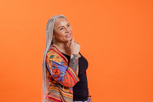 A front-view waist-up portrait shot of a mature woman with braided hair smiling. She is looking away from the camera with her had on her chin, while thinking. She is standing in front of an orange coloured background while wearing casual clothing.