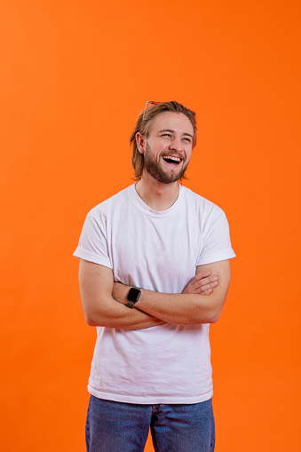 A front-view three quarter length portrait shot of a young man laughing and looking away the camera. He is standing in front of an orange coloured background while wearing casual clothing, he has his arms crossed.