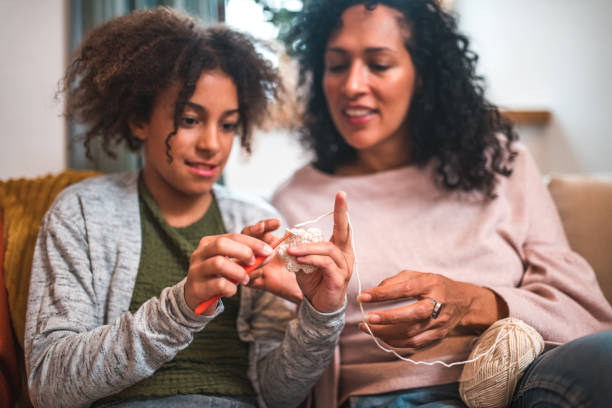 Smiling girl with an afro hairstyle learning how to crochet and knitting. With her Hispanic mother as a guiding hand, she discovers the joy of crafty hobbies.