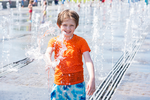 happy european boy 9 years old in a t-shirt and shorts plays in the fountain, touches the water jets and splashes with water on a sunny day outside